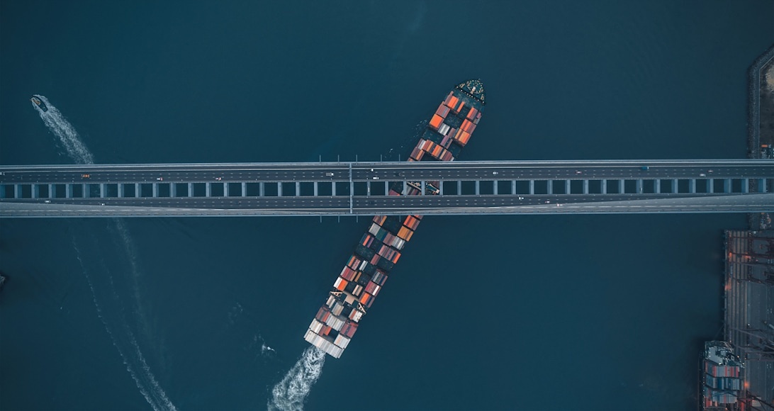 Ett stort containerfartyg fullastat med färgglada containrar passerar under en bro, vilket visar på den globala maritima logistikens omfattning.