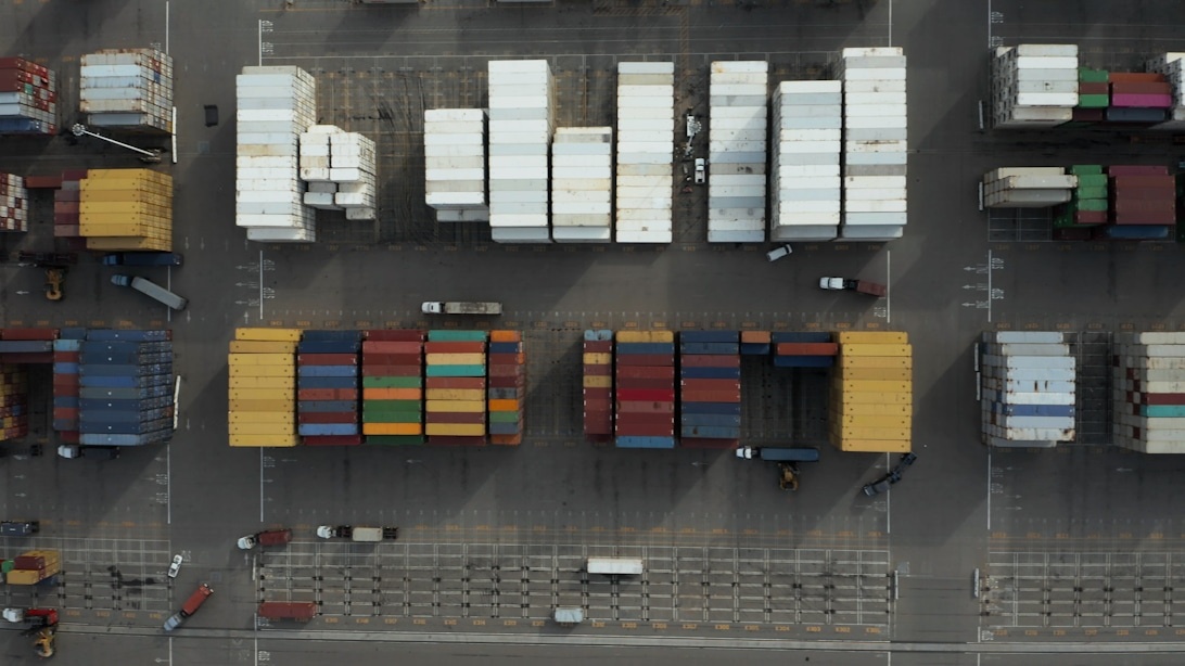 Flygfoto över en containerhamn med prydligt organiserade rader av containrar, lastbilar och annan logistisk infrastruktur.