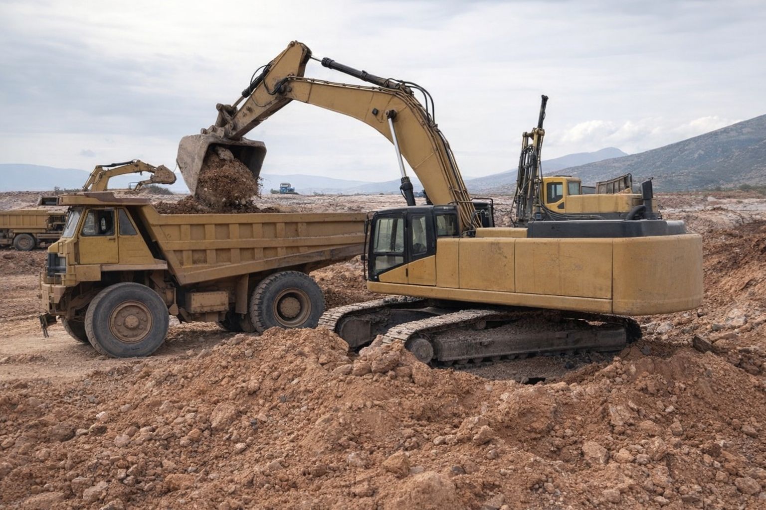 Gul grävmaskin lastar jord på en dumper vid en byggarbetsplats under en molnig himmel.